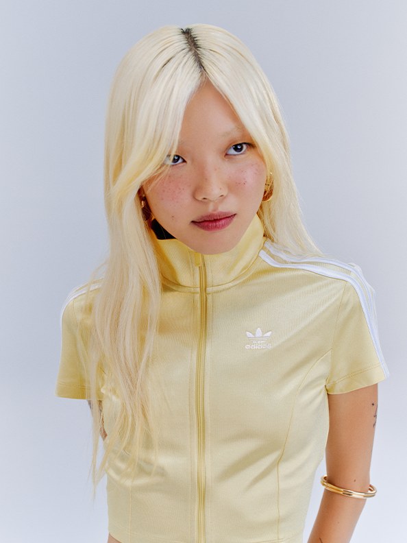 A young girl with blonde hair wearing a yellow jacket stands against a plain white background.