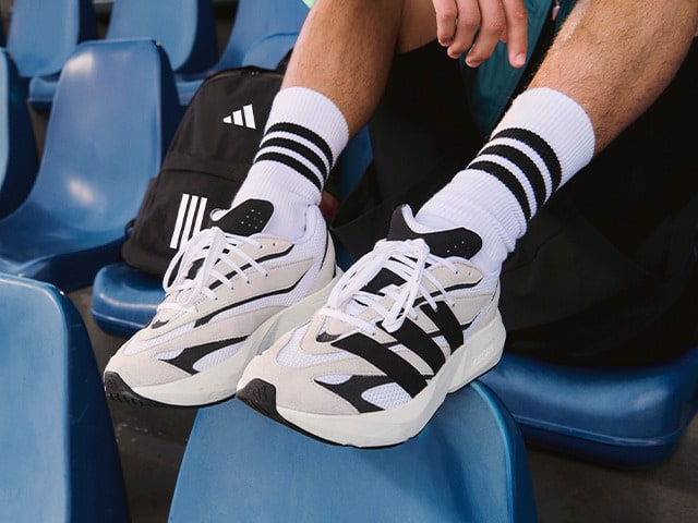 The image shows a pair of white and black athletic shoes with distinctive Adidas stripes on the sides, resting on a blue stadium seat.