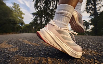 A close-up view of a person's feet wearing white and pink athletic shoes on a gravel path surrounded by trees.