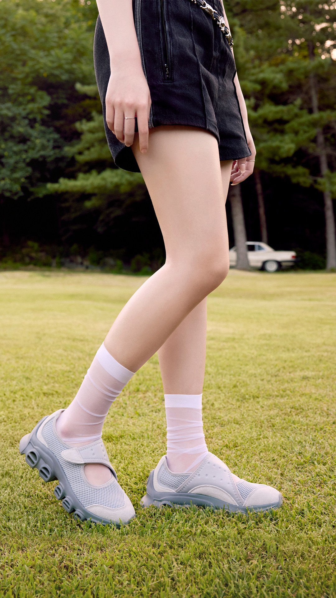 The image shows a person's lower body wearing black shorts and gray athletic shoes standing on a grassy field, with a blurred car in the background.