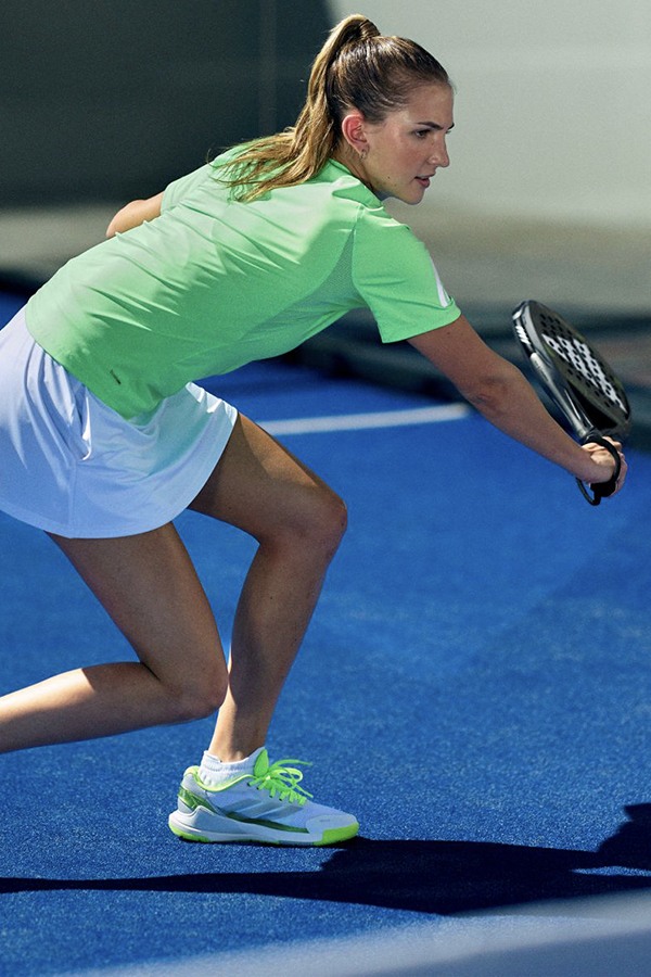 A female athlete in a bright green shirt and shorts is crouched on a blue tennis court, preparing to make a move.