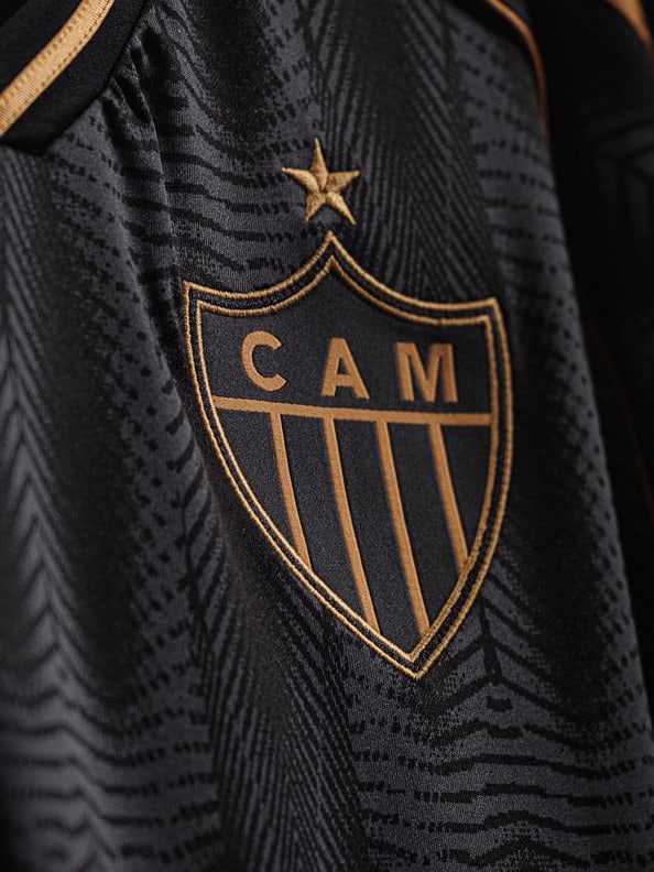 The image shows the logo of the Brazilian football club Atlético Mineiro, featuring a shield-shaped crest with the club's initials "CAM" and a star above it, set against a black and gold patterned background.