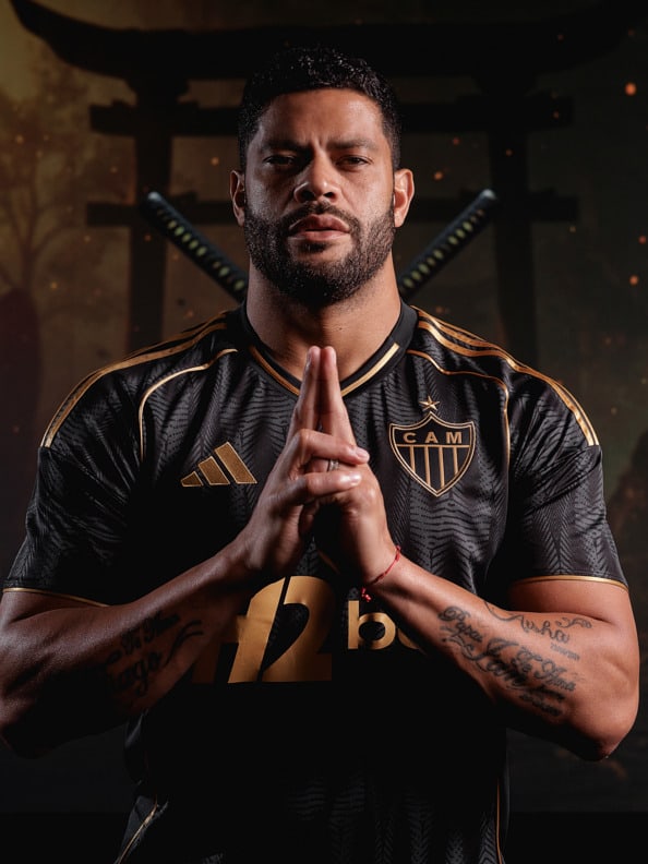 The image shows a man wearing a black and gold soccer jersey, with his hands clasped in a prayer-like gesture, against a dark background.