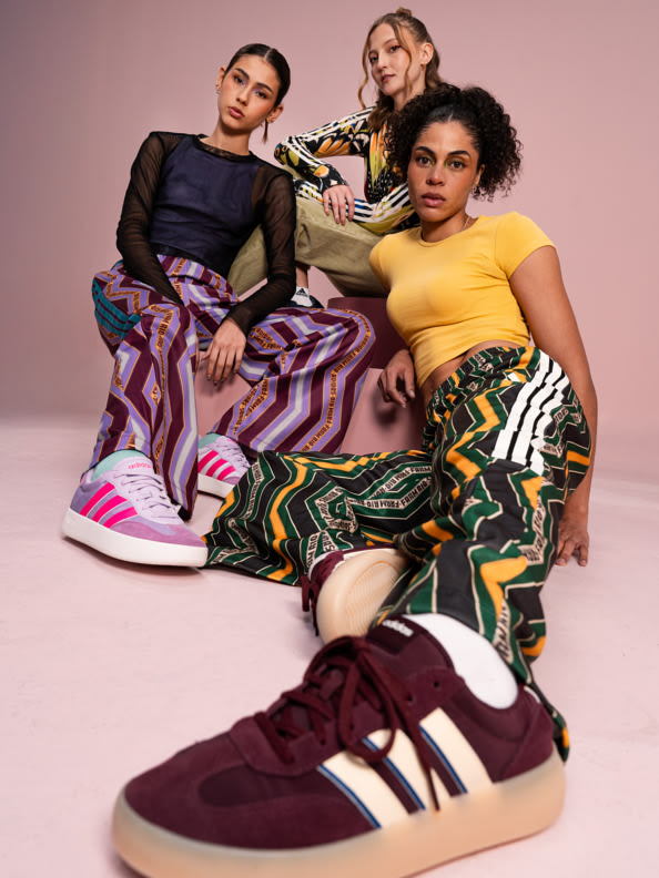 Three individuals, two women and one man, are seated on the floor in a studio setting with a pink background. They are wearing vibrant, patterned clothing and sneakers, creating a visually striking and stylish composition.
