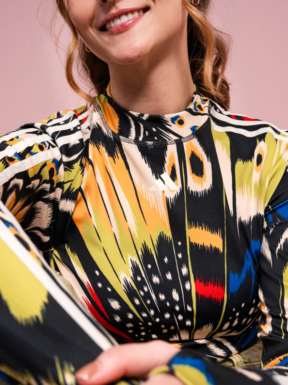 A smiling young woman wearing a vibrant, patterned dress stands against a plain pink background.