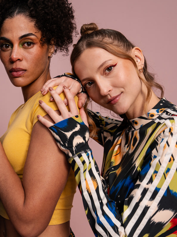 Two young women with diverse appearances and styles posing together against a plain pink background.
