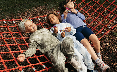 The image shows three young people, two women and one man, relaxing and enjoying themselves on a red climbing net in an outdoor setting with greenery in the background.