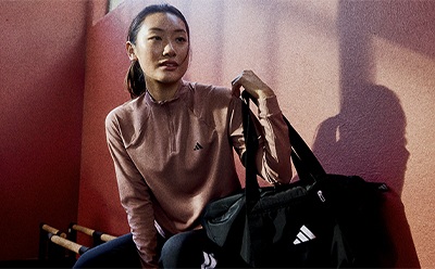 A young woman in a pink sweater stands in front of a red wall, holding a black sports bag.
