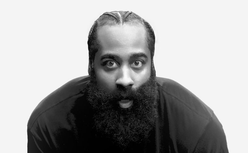 A close-up black and white portrait of a man with a long, thick beard and intense, wide-eyed expression, set against a plain, light-colored background.