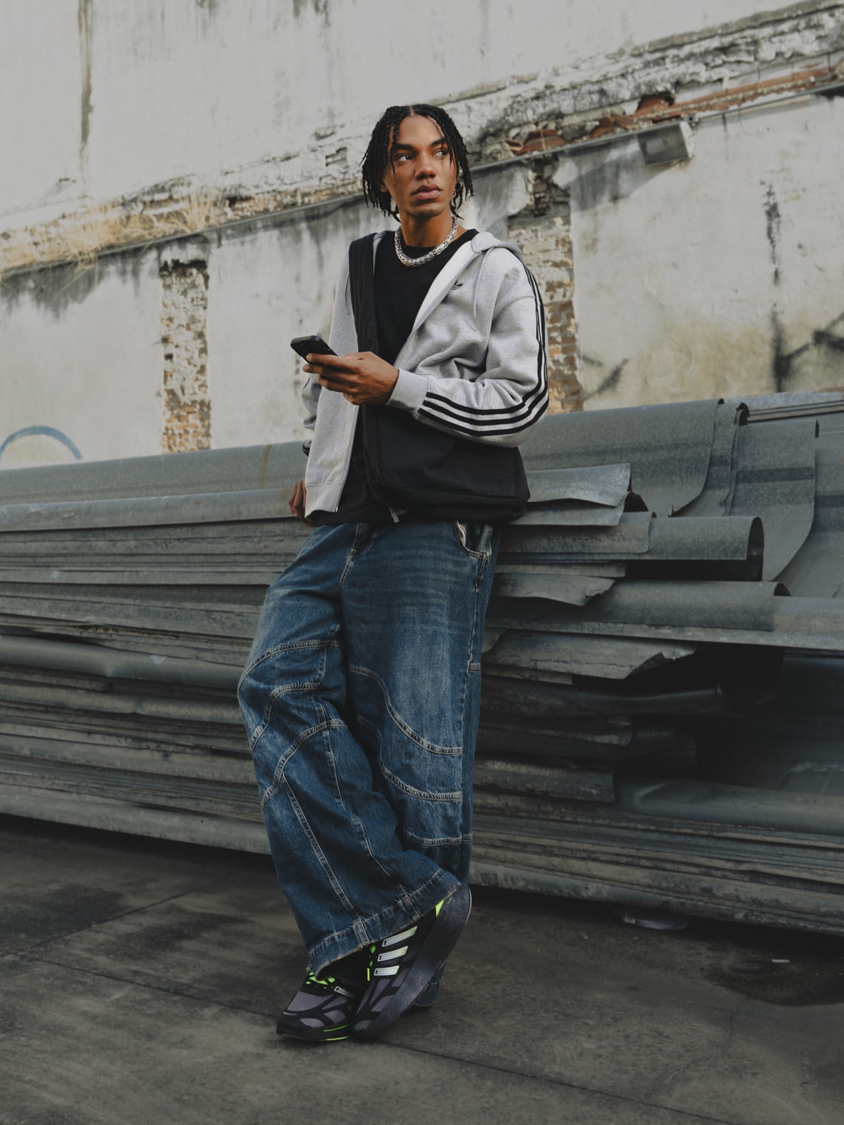 A young person with dreadlocks wearing a gray sweatshirt and jeans stands in front of a dilapidated wall, holding a mobile device.