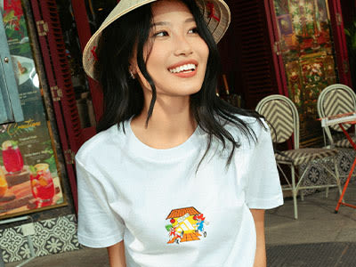 A young woman with long dark hair is smiling and wearing a white t-shirt with a colorful graphic design, standing in front of a patterned backdrop with a red door.