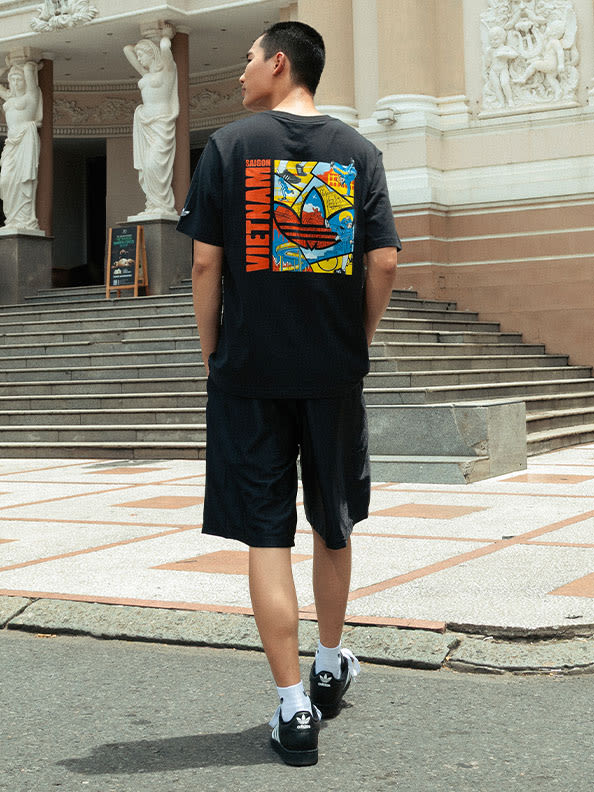 The image shows a person wearing a black t-shirt with a colorful graphic design on the back, standing on the steps of a building with statues in the background.