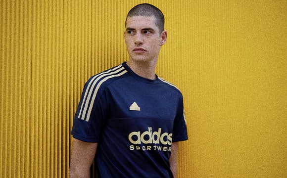A young man wearing a navy blue Adidas sportswear jersey stands against a vibrant yellow background.