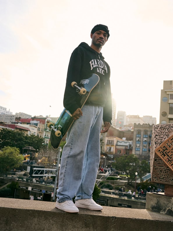 A person wearing a green sweatshirt and holding a skateboard stands on a platform overlooking a cityscape in the background.