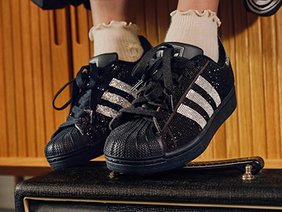 Op de afbeelding prijkt een paar zwarte en witte adidas Superstar sneakers die rusten op een vintage versterker, met de benen en voeten van de drager zichtbaar op de voorgrond.