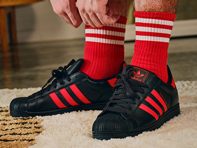 Bilden visar en persons underkropp iklädd röda och vita randiga strumpor och svarta och röda adidassneakers, som står på en beige matta.