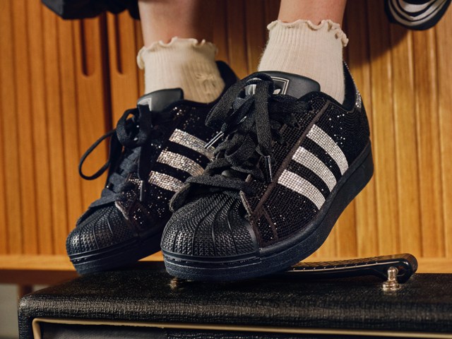 A pair of black and white sneakers with sparkling accents sits on a wooden surface, with a curtain-like backdrop in the background.