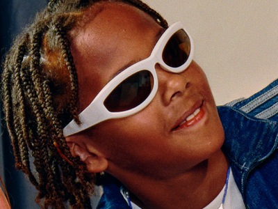 A young person with braided hair wearing white sunglasses and a denim jacket, appearing to be smiling and looking off to the side.