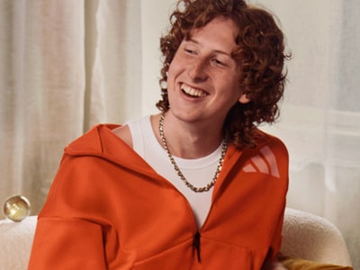 En leende individ med lockigt hår iklädd en orange jacka med huva sitter i en stol mot en ljus bakgrund.