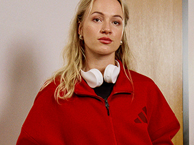 Een jonge vrouw met blond haar die een felrood jack draagt staat tegen een effen muur achtergrond.