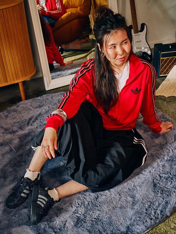 Een jonge vrouw met lang donker haar zit op een grijs tapijt in een kamer gevuld met verschillende objecten, waaronder een gitaar en versterker.