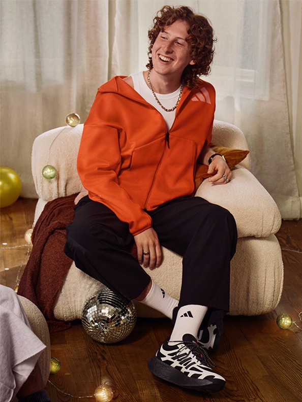 En person med lockigt hår som bär en orange hoodie sitter i en soffa, omgiven av olika föremål inklusive en discoboll och några äpplen.