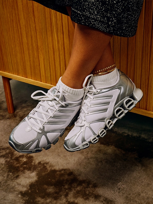 De afbeelding toont een paar grijze en witte sportschoenen met een opvallend design, rustend op een houten vloer tegen een houten achtergrond.