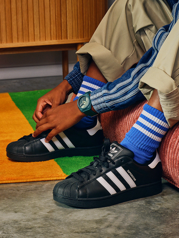 Bilden visar en persons underkropp iklädd ett par sneakers från adidas, blå och vita randiga strumpor och en beige och blå outfit som sitter på en färgglad matta i vad som verkar vara en inomhusmiljö med träpaneler på väggarna.