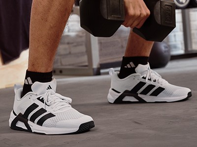 Bilden visar en persons underkropp iklädd vita och svarta sportskor, som står på ett betonggolv i vad som verkar vara ett gym eller fitnesscenter.