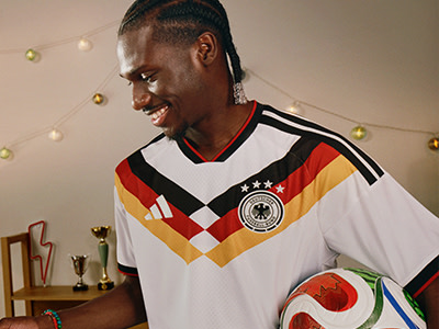 En leende ung man som bär en vit, röd och svart fotbollströja står i ett rum med hyllor som visar olika troféer och dekorationer.