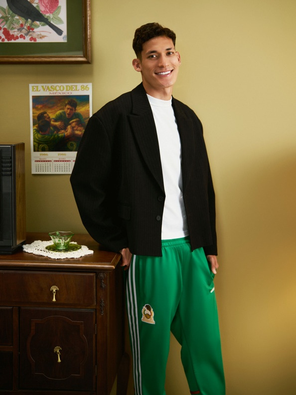 The image shows a young man wearing a black jacket and green pants, standing in front of a wooden dresser with a framed artwork on the wall behind him.