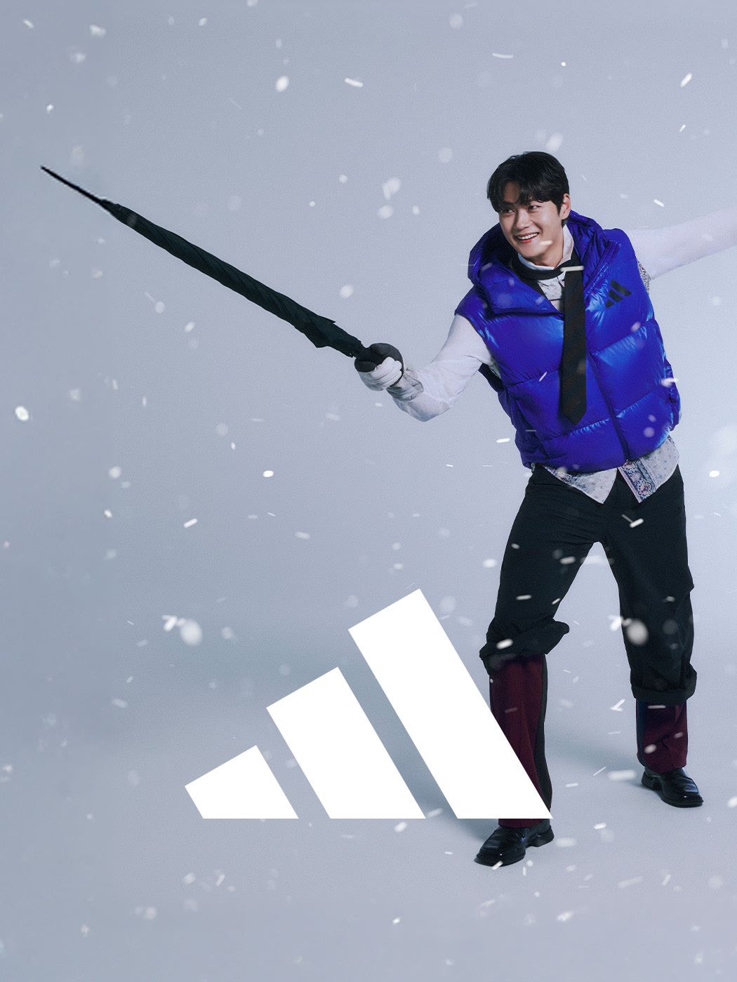 A person in a blue jacket is holding a large white Adidas logo in the middle of a snowy landscape.