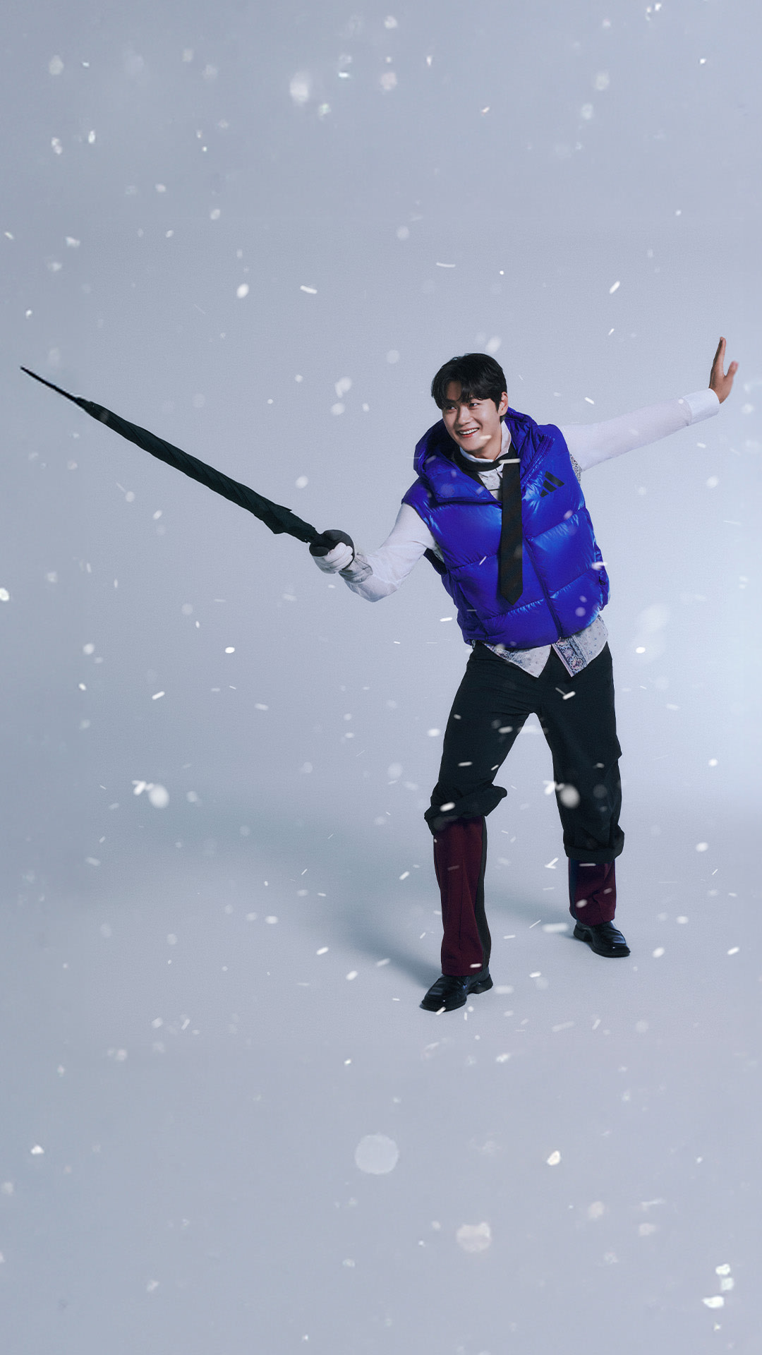 A person in a blue jacket and winter gear stands in a snowy landscape, holding a long pole and making a gesture with their free hand.