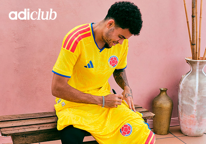 A man wearing a yellow soccer jersey is sitting on a wooden bench in front of a pink wall, with decorative vases and other items visible in the background.