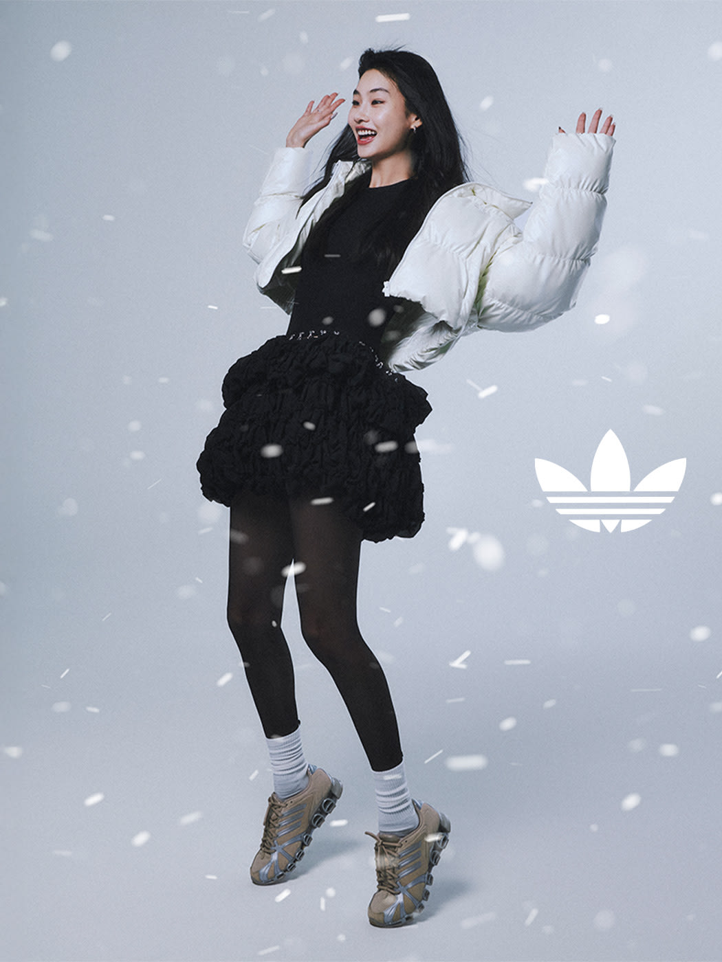 A young woman in a black outfit with a puffy white jacket stands in a snowy setting, her arms raised as if dancing or celebrating.