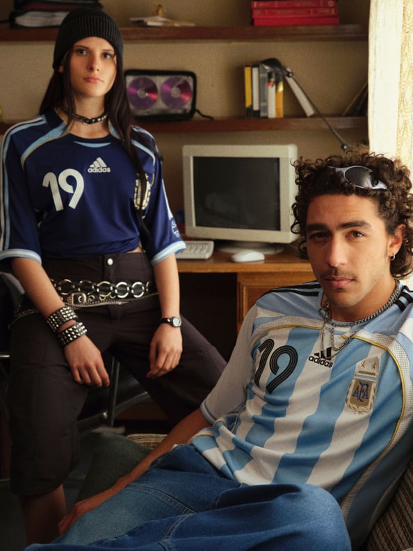 The image shows a young woman wearing a navy blue sports jersey with the number 19 and a young man wearing an Argentinian national team jersey, both sitting in a room with shelves and a computer monitor in the background.