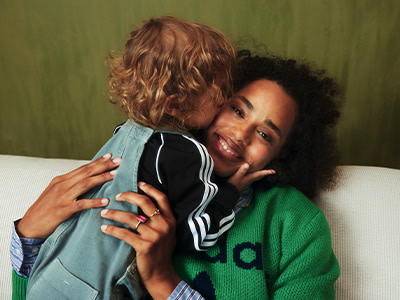 Bildet viser en varm omfavnelse mellom to individer, én med krøllete hår og den andre med en livlig grønn genser, satt mot en vanlig grønn bakgrunn.