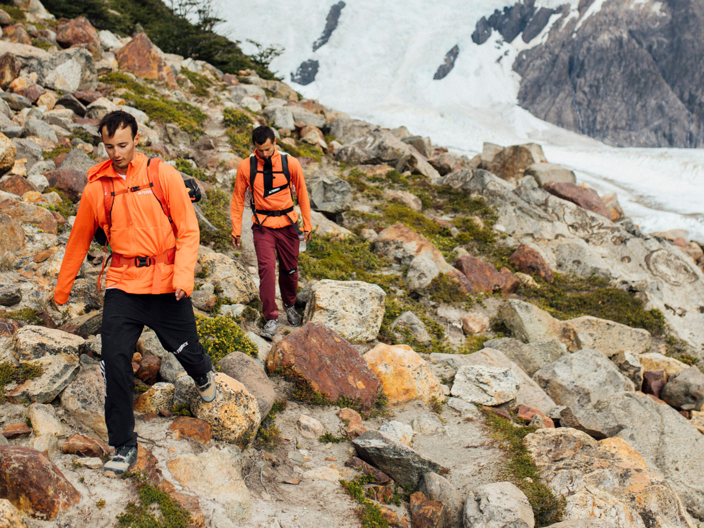 Two hikers in bright orange and red jackets stand on a rocky, mountainous terrain with snow-capped peaks in the background.