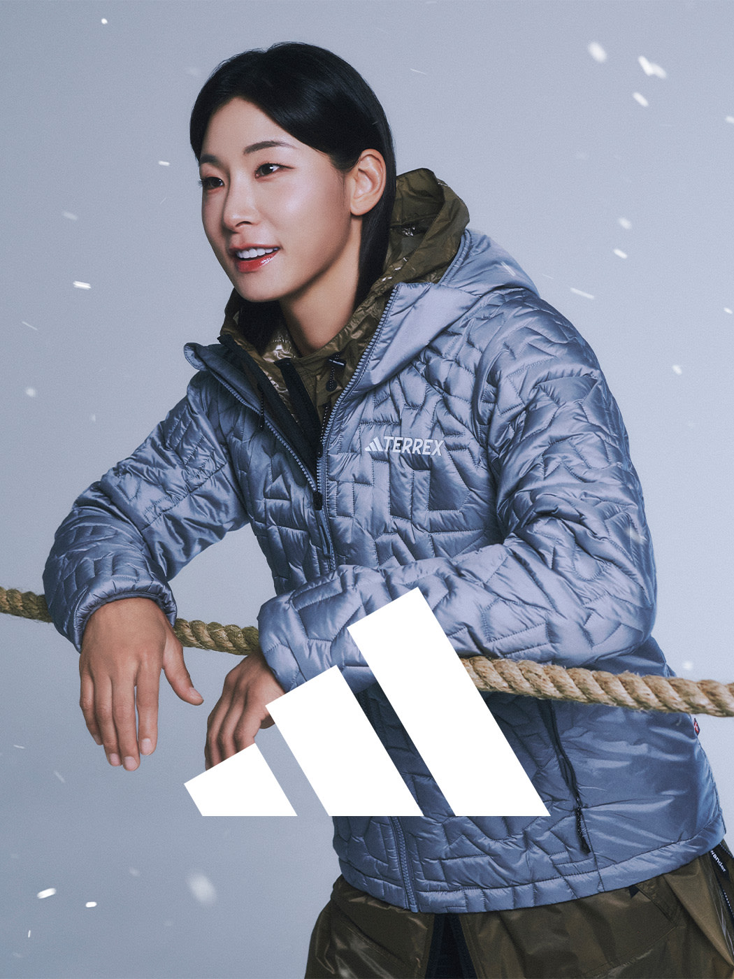 A person wearing a gray and olive green winter jacket stands in a snowy outdoor setting, with a white background and falling snow visible.