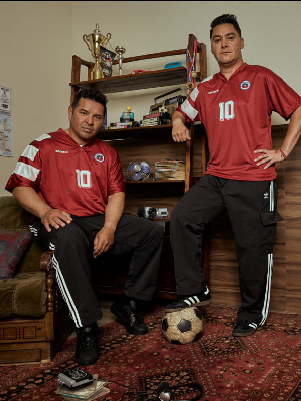 Two men wearing red soccer jerseys with the number 10 are sitting on a couch in a room with various sports-related items and trophies displayed on shelves in the background.