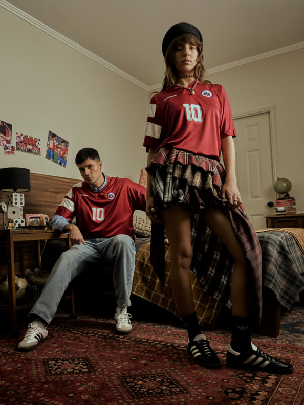 The image shows two individuals, a man and a woman, wearing matching red sports jerseys with the number 10 on them, sitting on a rug in what appears to be a room with various personal items and decorations visible in the background.