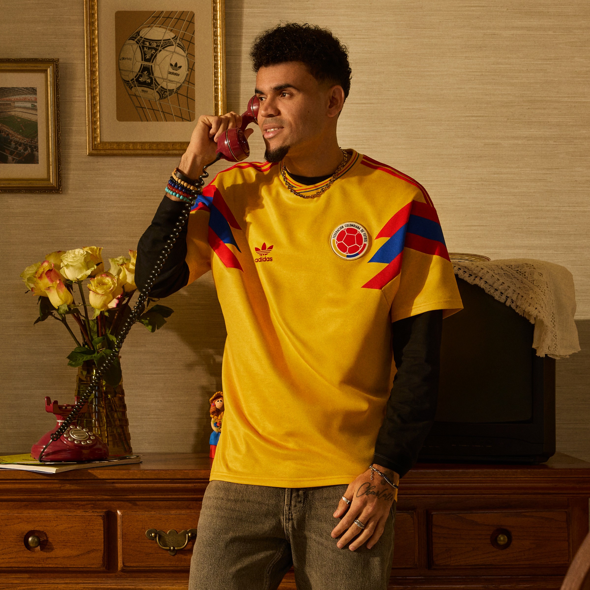 A young man wearing a vibrant yellow and red soccer jersey is sitting on a couch in a room with framed artwork and a vase of flowers on a wooden dresser behind him.