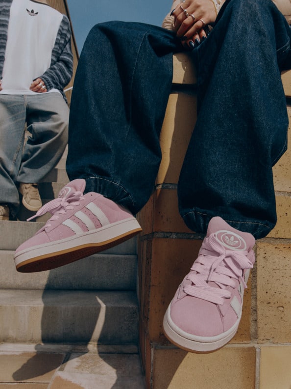 The image shows a person's legs and feet, with the person wearing a pair of pink Adidas sneakers and dark blue jeans. The background appears to be a wooden surface or floor.
