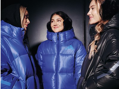 Three women wearing colorful puffy jackets stand against a dark background.