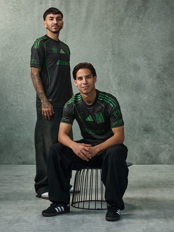 Two individuals wearing matching green and black athletic jerseys are posing in a studio setting with a plain gray background.