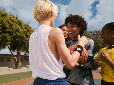 A group of young people, including a blonde woman and a man with curly hair, are interacting and playing basketball on an outdoor basketball court surrounded by trees and a cloudy sky.