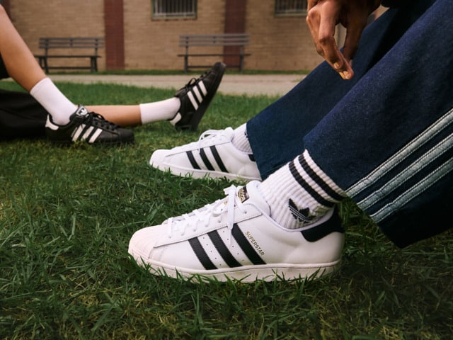 The image shows a pair of white and black Adidas sneakers on a grassy field, with a brick building visible in the background.