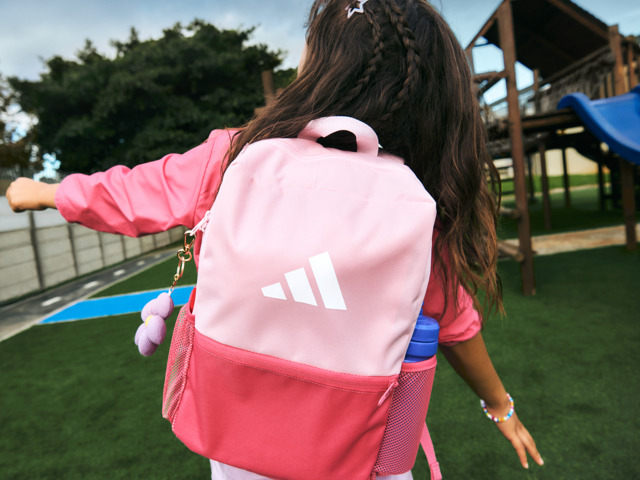 A young person wearing a pink and white backpack stands in a grassy outdoor area with playground equipment visible in the background.