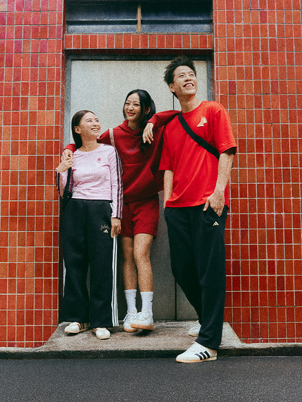 Three young people, two women and one man, are standing together in front of a red brick wall, smiling and embracing each other. They appear to be wearing casual, coordinated outfits in shades of red and black.