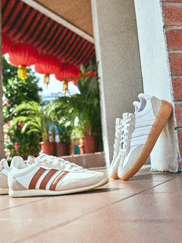 A pair of white and red sneakers sits on a wooden floor, with a red and white striped canopy and lush greenery visible in the background.